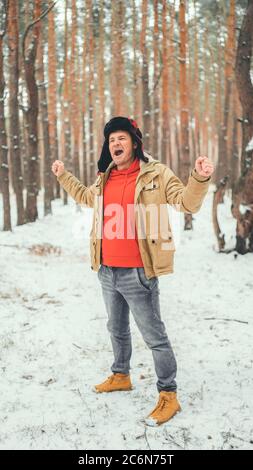 Schöner Mann steht und singt im Wald in der Wintersaison. Junger Mann trägt beige Jacke über roten Hoodie und Ohrklappe Hut mit seinen Armen Stockfoto