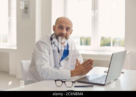 Der Praktizierende Arzt senier sitzt mit einem Laptop am Arbeitsplatz im Büro der Klinik an einem Tisch. Stockfoto