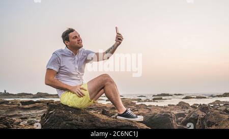 Entspannter Mann sitzt auf Stein und Selfie auf dem Smartphone am Ufer. Seitenansicht des angenehmen Mannes, der Zeit verbringt, den Urlaub zu genießen und zu fotografieren Stockfoto