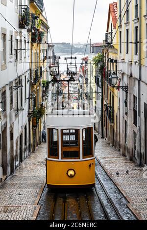 Traditionelle gelbe Straßenbahn in einer engen Straße in Lissabon an einem regnerischen Wintertag, mit dem Fluss Tejo im Hintergrund entkokend. Stockfoto