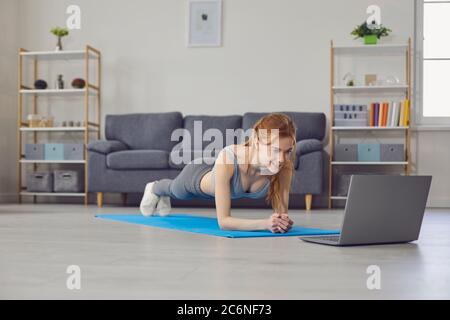 Online-Heimsport. Hübsches Mädchen, das im Wohnzimmer mit einem Laptop ausarbeitet. Frau macht Ellenbogen Plank Übung Stockfoto