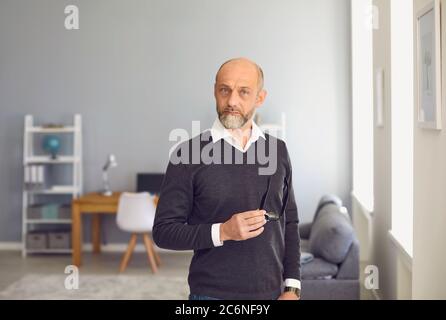 Porträt eines älteren Mannes, der zu Hause auf die Kamera schaut. Serious reifen Mann stehend mit Brille in der Hand drinnen Stockfoto