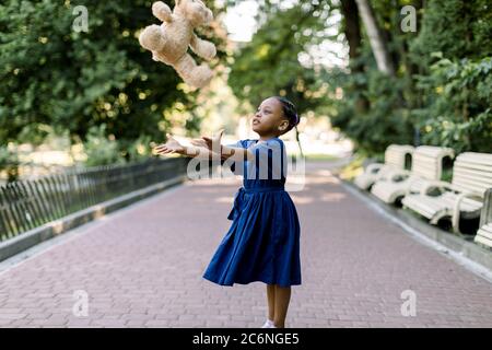 Portrait von glücklichen kleinen afrikanischen Mädchen Kind im grünen Stadtpark werfen Teddybär Spielzeug, fliegen in der Luft. Lächelnd dunkelhäutig Mädchen in blau Stockfoto