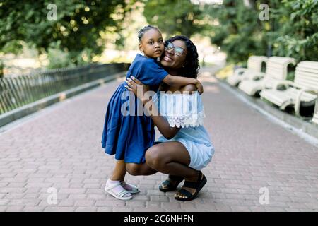 Full length Portrait von lächelnden jungen glücklich afrikanischen Mutter umarmt ihre kleine Tochter mit Zärtlichkeit und Liebe im Park stehen. Mutter und Stockfoto