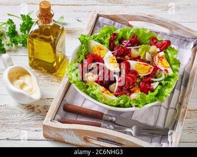 Nahaufnahme von Baby-Oktopus-Salat mit hart gekochten Eiern, Kapern, marinierte Zwiebel auf Salatblättern in einer Schüssel auf einem Tablett, auf einem rustikalen Holztisch mit Oli Stockfoto