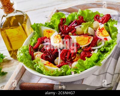 Nahaufnahme von Baby-Oktopus-Salat mit hart gekochten Eiern, Kapern, marinierte Zwiebel auf Salatblättern in einer Schüssel auf einem Tablett, auf einem rustikalen Holztisch mit Oli Stockfoto