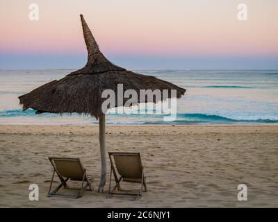 Atemberaubender Sonnenaufgang mit einem traditionellen Strohschirm und zwei Liegestühlen mit Blick auf die friedliche Küste des Indischen Ozeans, Anakao, Madagaskar Stockfoto