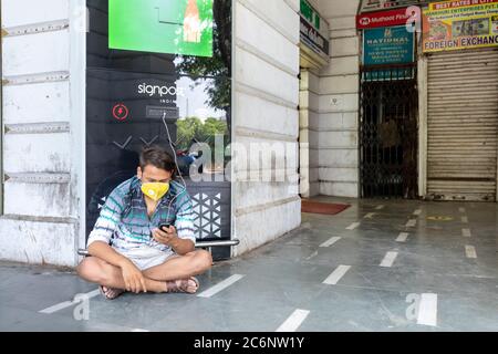 Ein Mann sitzt auf dem Boden und schaut auf sein Telefon, während er eine Corona Schutzmaske in Connaught Place trägt Stockfoto