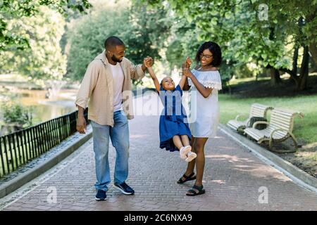 Glückliche afrikanische Familie im Park im Sommer sonnigen Abend. Mama, Papa und glückliche Tochter gehen bei Sonnenuntergang, Eltern halten die Hände des kleinen Mädchens Stockfoto