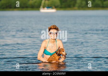 Reife kaukasische Frau hilft ihrem kleinen alten Dachshund Hund im Wasser zu schwimmen. Verbringen Sie die Sommerzeit mit einem Hund auf dem Fluss. Urlaubstag am See Stockfoto