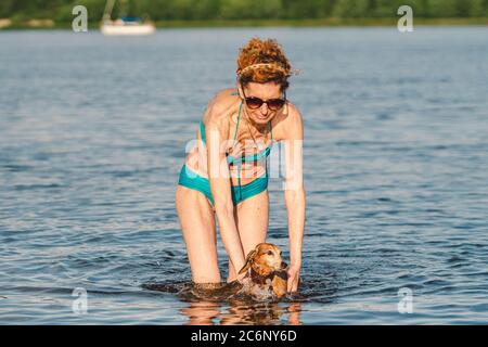 Reife kaukasische Frau hilft ihrem kleinen alten Dachshund Hund im Wasser zu schwimmen. Verbringen Sie die Sommerzeit mit einem Hund auf dem Fluss. Urlaubstag am See Stockfoto