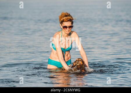 Reife kaukasische Frau hilft ihrem kleinen alten Dachshund Hund im Wasser zu schwimmen. Verbringen Sie die Sommerzeit mit einem Hund auf dem Fluss. Urlaubstag am See Stockfoto