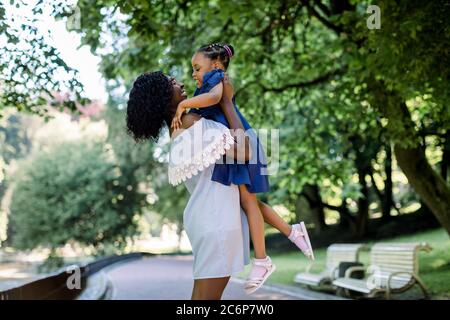 Junge schöne afrikanische Frau Mutter in hellblauem Kleid spielt mit ihrer kleinen niedlichen Tochter im Park, Spaß haben und das Baby heben. Glücklich Stockfoto