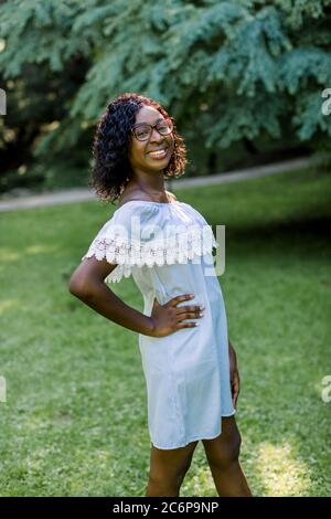 Outdoor-Porträt der jungen schönen afrikanischen Mädchen in blauem Kleid, mit charmanten Lächeln, kurze dunkle lockige Haare, genießen ihren Spaziergang in schönen Park Stockfoto