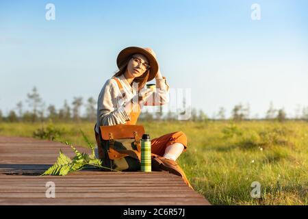 Botanistin mit Rucksack auf Holzweg durch Moor Sumpf im Wildlife National Park sitzen. Naturforscher auf der Promenade ruhen, Tee trinken, e Stockfoto