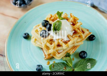 Frisch gebackene hausgemachte klassische belgische Waffeln mit Eiscreme, frischen Blaubeeren und Minze auf Holzhintergrund, Blick von oben. Herzhafte Waffeln Stockfoto