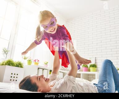 Mutter und ihr Kind spielen zusammen. Mädchen in einem Superhelden Kostüm. Das Kind hat Spaß und springt auf dem Bett. Stockfoto