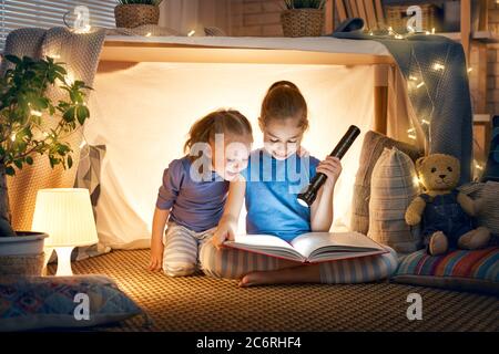 Zwei niedliche kleine Kinder lesen ein Buch mit Taschenlampen im Zelt. Glückliche Mädchen spielen zu Hause. Lustige, nette Kinder, die Spaß im Zimmer haben. Stockfoto