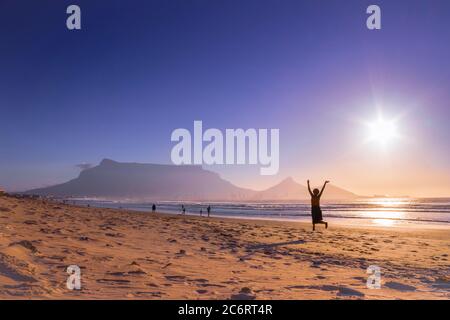 Zwei afrikanische Frauen tanzen am Strand bei Sonnenuntergang, mit Tafelberg und Kapstadt im Hintergrund, Milnerton Beach, Kapstadt, Südafrika Stockfoto