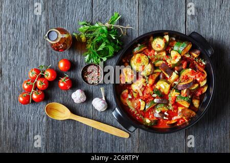 Gemüseeintopf, Auberginen, Zwiebel, Zucchini mit Tomatensauce, Knoblauch und Kräuter in einer Auflaufform auf einem Holztisch mit Zutaten, Landschaftsansicht von Stockfoto