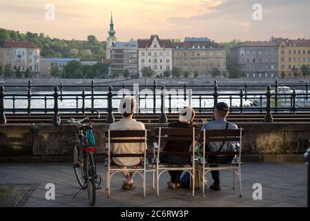 Drei Personen sitzen auf Stühlen an der Donaupromenade genießen Budapest Aussicht. Rückansicht Stockfoto