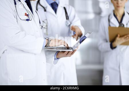 Eine Gruppe unbekannter Ärzte, Männer und eine Frau mit Stethoskopen diskutieren medizinische Untersuchungsresoults, mit einem Laptop, während sie im Krankenhaus stehen Stockfoto