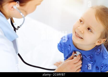 Arzt untersucht ein Kind mit Stethoskop in der sonnigen Klinik. Glücklich lächelnd Mädchen Patient in blauem Kleid gekleidet ist bei der üblichen medizinischen Inspektion Stockfoto