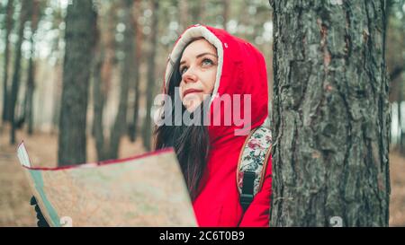 Eine Reisende Frau mit Karte im Wald. Ein Porträt der hübschen Frau mit Rucksack, die bei kaltem Wetter in der Nähe eines Baumes steht. Stockfoto