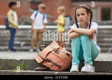 Full length Portrait of Sad Schulmädchen Blick auf Kamera, während auf Treppen im Freien mit Gruppe von Kindern im Hintergrund sitzen, kopieren Raum Stockfoto