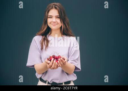 Junge stilvolle trendige Frau isoliert über graublauen Hintergrund. Attraktive schöne Mädchen hält rote reife Kirschen. Allein auf der Kamera bei Dunkelheit wal posieren Stockfoto