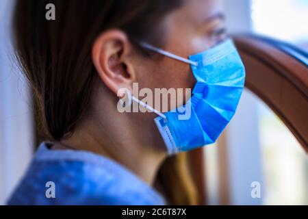 Frau, die am Fenster mit einer Maske auf ihrem Gesicht steht. Bleiben Sie zu Hause. Selbstisolierung ist die einzige Chance, den Covid-19 zu stoppen. Aufenthalt zu Hause Quarantäne Co Stockfoto