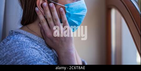 Frau, die am Fenster mit einer Maske auf ihrem Gesicht steht. Bleiben Sie zu Hause. Selbstisolierung ist die einzige Chance, den Covid-19 zu stoppen. Aufenthalt zu Hause Quarantäne Co Stockfoto