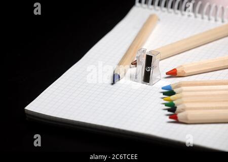 Weißes Notizbuch, Farbstifte und Bleistiftspitzer auf einem Tisch vor schwarzem Hintergrund. Vorbereitung auf eine Schule. Zurück zur Schule. Stockfoto