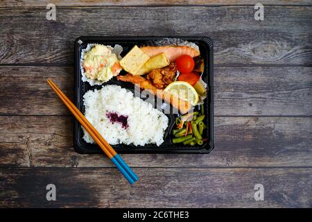 Bunte japanische „Makunouchi“ Bento-Box mit Lachs, gebratenen Garnelen, Kartoffelsalat, Tofu, gegrilltem Hähnchen, Reis und Gemüse. Rustikaler dunkler Holztisch. Stockfoto