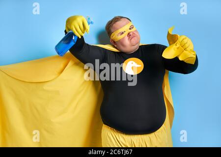 Portrait von lustigen fröhlichen fetten Mann mit Waschmittel in den Händen, ist der Mann bereit zu reinigen, trägt gelbe Kostüm, Handschuhe und Maske von Superhelden. Isoliert blau Stockfoto