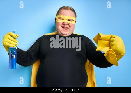 Portrait von lustigen fröhlichen fetten Mann mit Waschmittel in den Händen, ist der Mann bereit zu reinigen, trägt gelbe Kostüm, Handschuhe und Maske von Superhelden. Isoliert blau Stockfoto