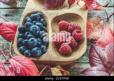 Wilde Beeren, viele Blaubeeren und Himbeeren in einem umweltfreundlichen Holzbecher auf einem Holztisch mit Herbstblättern. Stockfoto