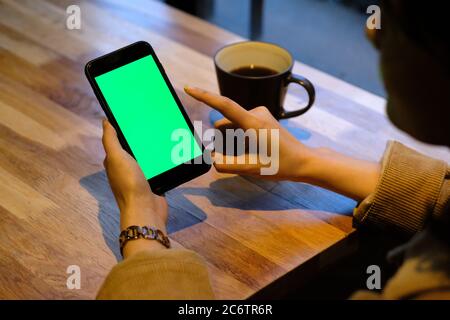 Über der Schulter eine Frau tippen grünen Bildschirm Smartphone im Café. Eine Tasse Kaffee auf einem Holztisch. Hintergrund verwischen Stockfoto