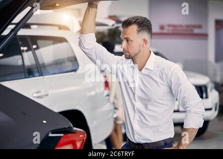 Handsome kaukasischen Kerl untersuchen den Kofferraum des Autos in Händler, er schaut in das Auto von hinten und denken, kaufen es oder nicht Stockfoto