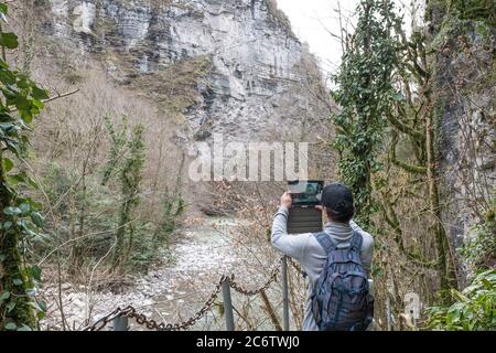 Im Winter ist Senior Mann mit Rucksack und in Baseball-Cap zu Fuß entlang Wanderweg. Er macht Fotos auf Tablette in Schlucht von Eibe und Buchsbaumhain von C Stockfoto