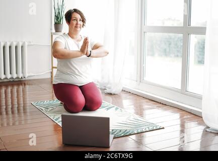 Erwachsene charmante Brünette Frau plus Größe Körper positive Praxis Yoga mit Laptop zu Hause Stockfoto