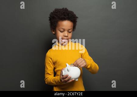 Niedliches schwarzes Kind, das in Sparschwein Euro-Münze setzt Stockfoto