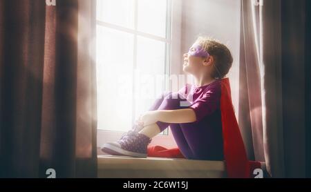 Kleines Mädchen spielt Superheld. Kind sitzt auf dem Fenster. Mädchen Power Konzept. Stockfoto