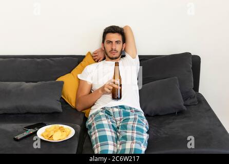 Ein junger Mann sitzt zu Hause auf der Couch und schaut fern. Er trinkt Bier und isst Chips. Stockfoto