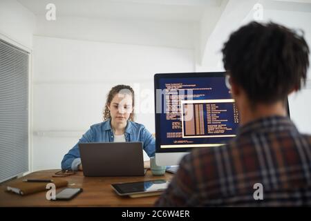 Rückansicht Portrait eines afroamerikanischen Mannes, der Code auf dem Computerbildschirm schreibt, während er mit einer Kollegin am Schreibtisch in der Softwareentwicklungsagentur arbeitet Stockfoto