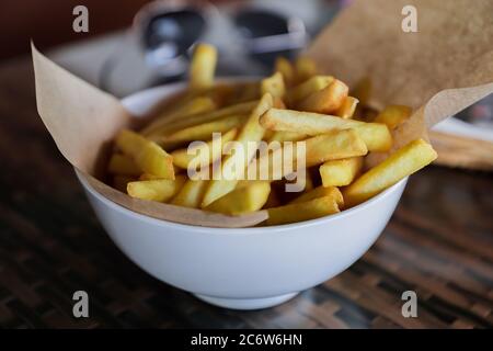 pommes Frites in einer weißen Schüssel serviert mit Bastelpapier auf dem Tisch, selektiver Fokus Stockfoto