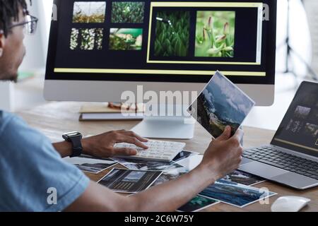 Rückansicht Porträt des jungen afroamerikanischen Mannes, der gedruckte Fotos hält, während er mit Bearbeitungssoftware über Computer arbeitet, während er am Schreibtisch in der Wohnung arbeitet Stockfoto