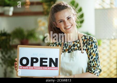 Eröffnung von Kleinunternehmen nach der Pandemie des Covid-19. Porträt der glücklichen modernen 40 Jahre alten kleinen Unternehmer Frau in Schürze zeigt offene Zeichen. Stockfoto