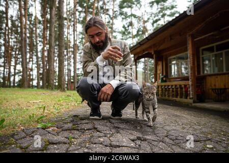 Ein Mann spielt mit einer grauen gestromten Katze. Ein Mann hält ein Glas in der Hand Stockfoto