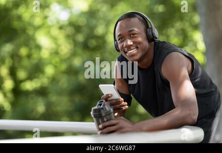 Lächelnder afrikanischer Sportler, der Wasser trinkt und Musik hört Stockfoto